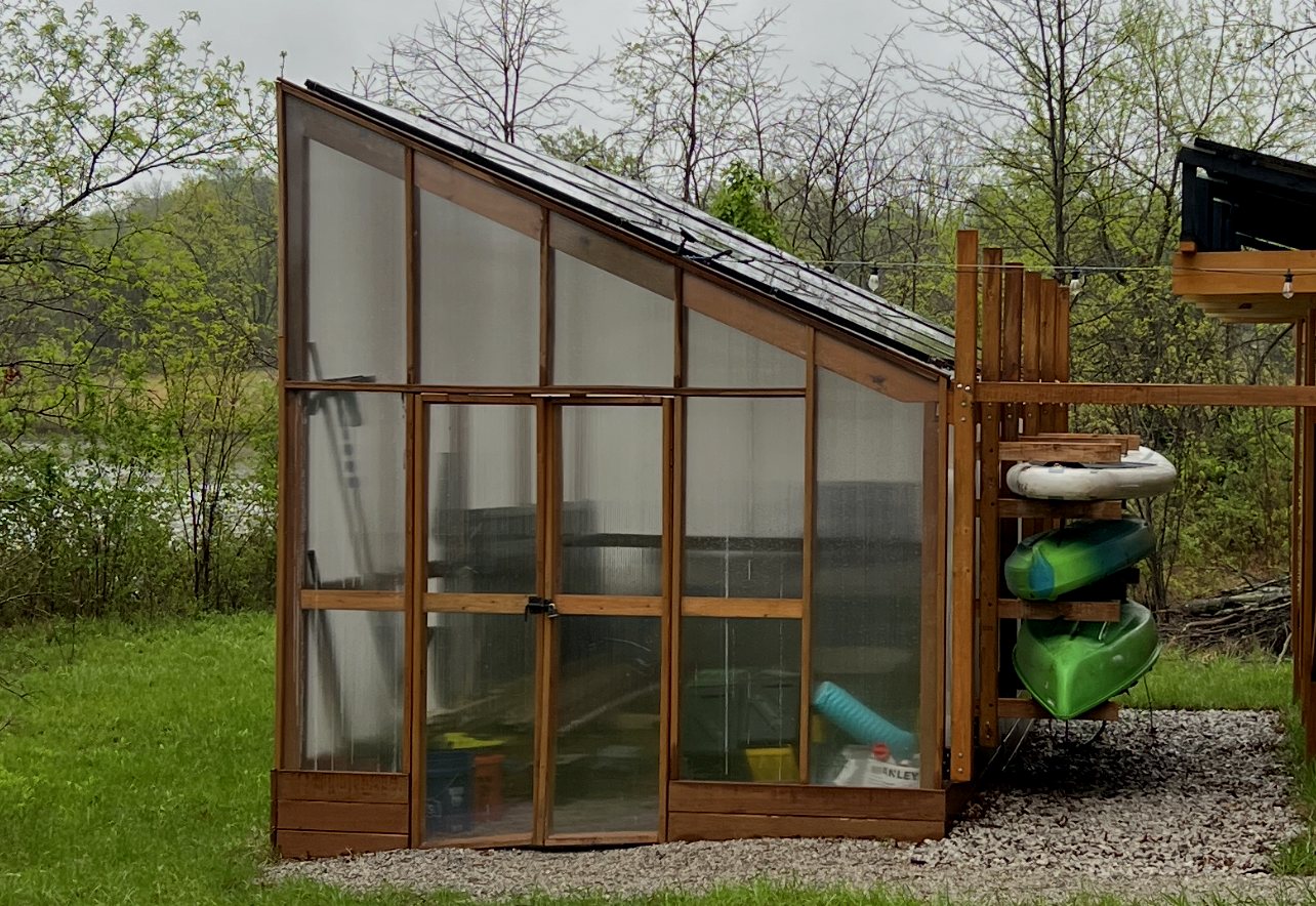 Cedar-framed lean-to greenhouse with solar panels on its sloping roof, next to a kayak rack